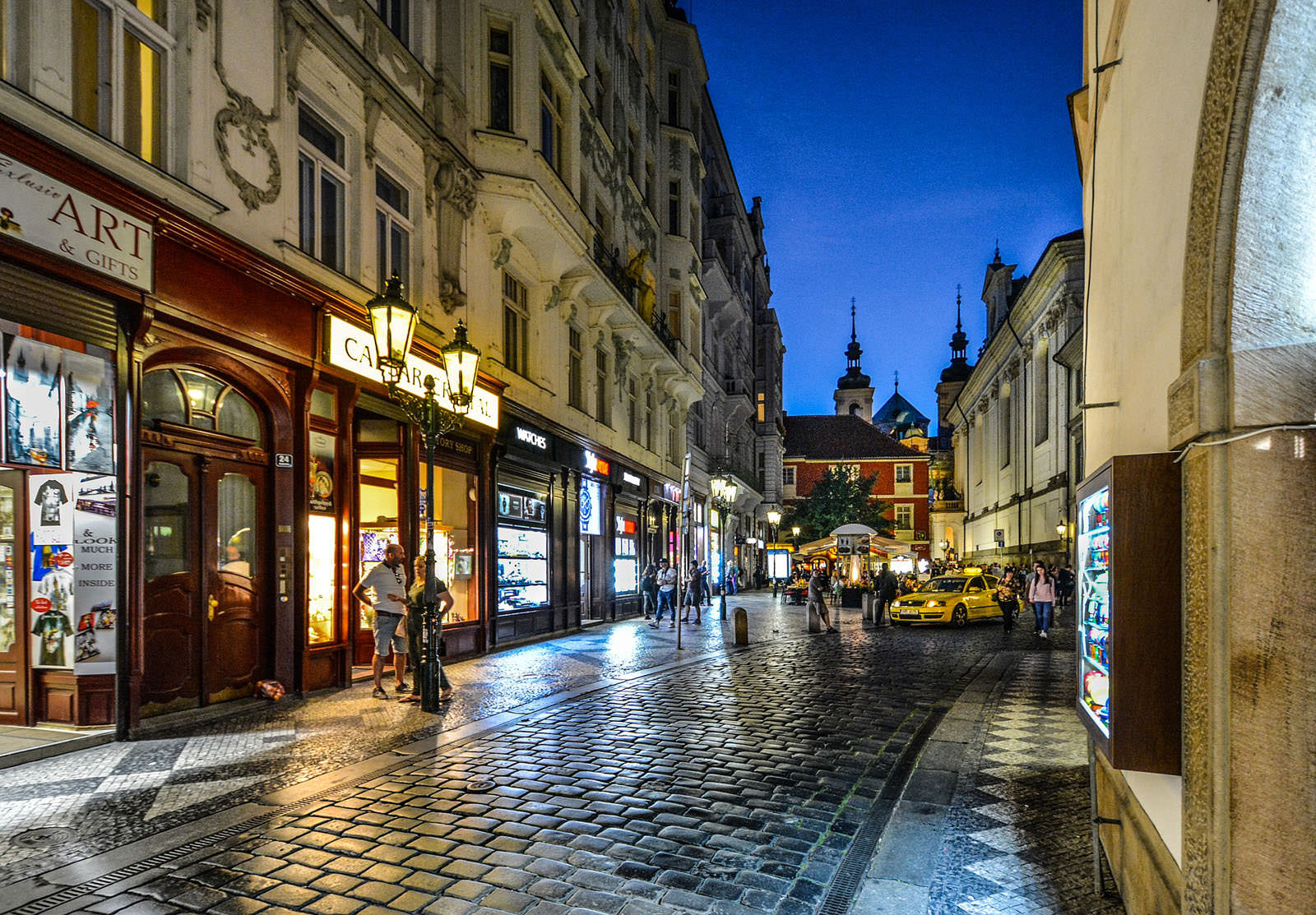 Old Town Prague street with souvenir shops, antique lamps, and taxi in front of Clementinum.