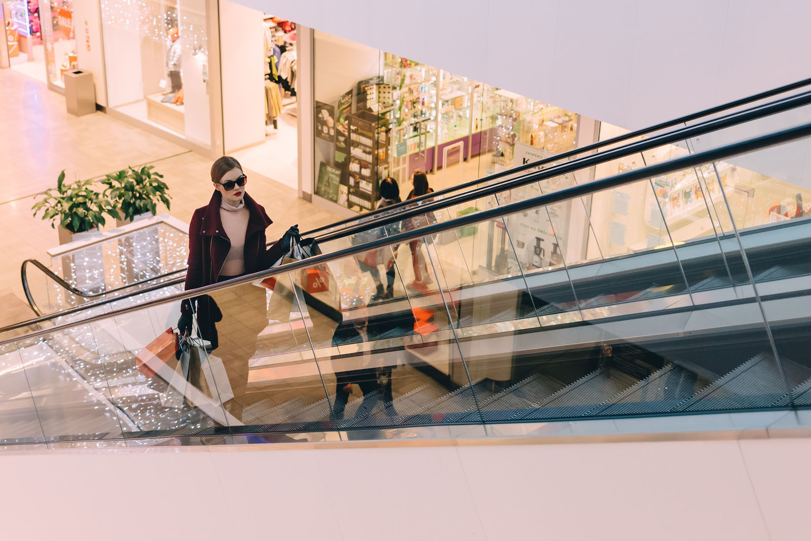 A woman riding an escalator in a shopping mall, holding shopping bags in both hands, with shop windows in the background.